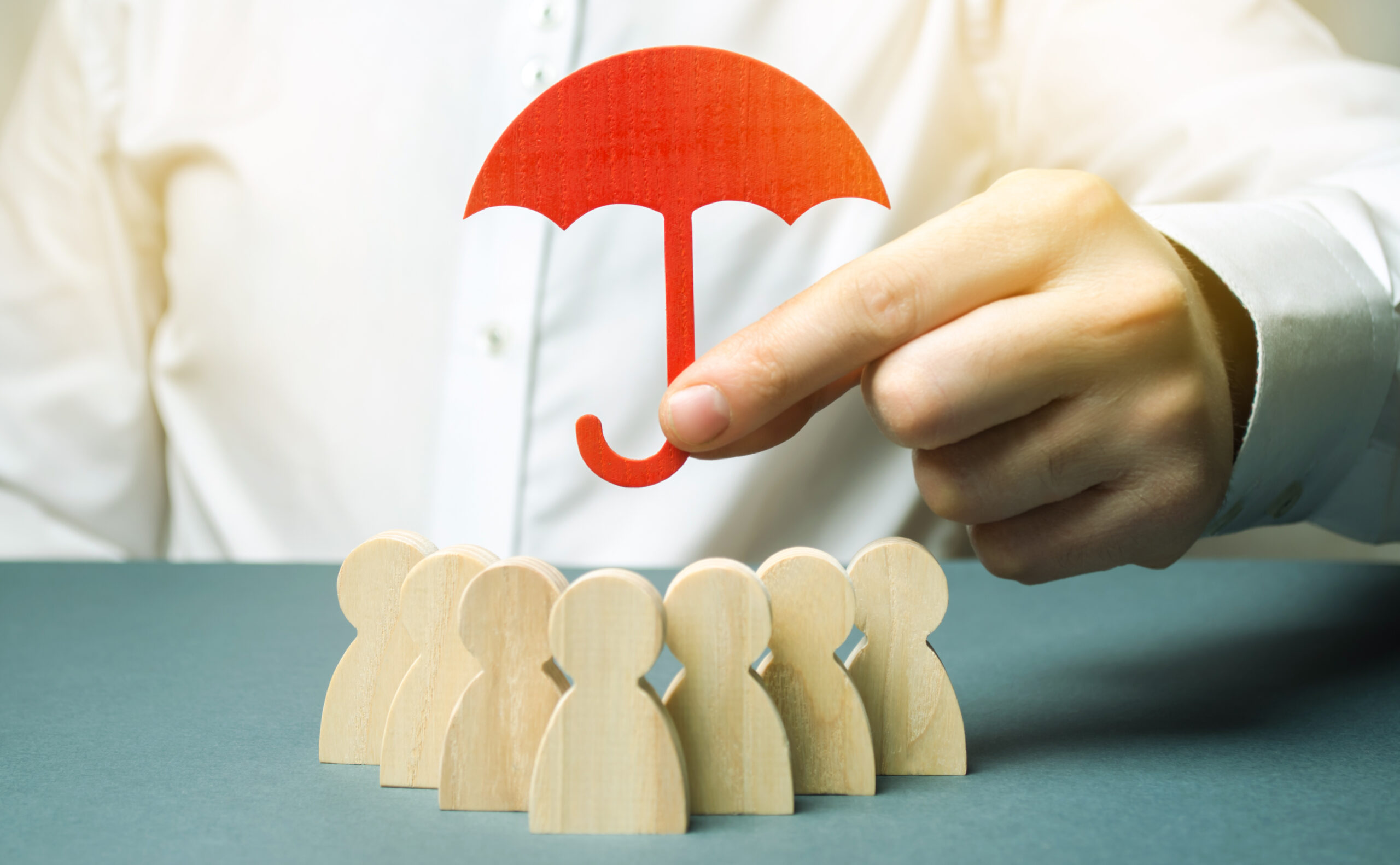 Boss holding a red umbrella and defending his team with a gesture of protection. Life insurance. Customer care, care for employees. Security and safety in a business team. Selective focus on voit une main d'humain, qui tien par deux doigts un petit parapluie rouge au dessus de bonhommes en bois